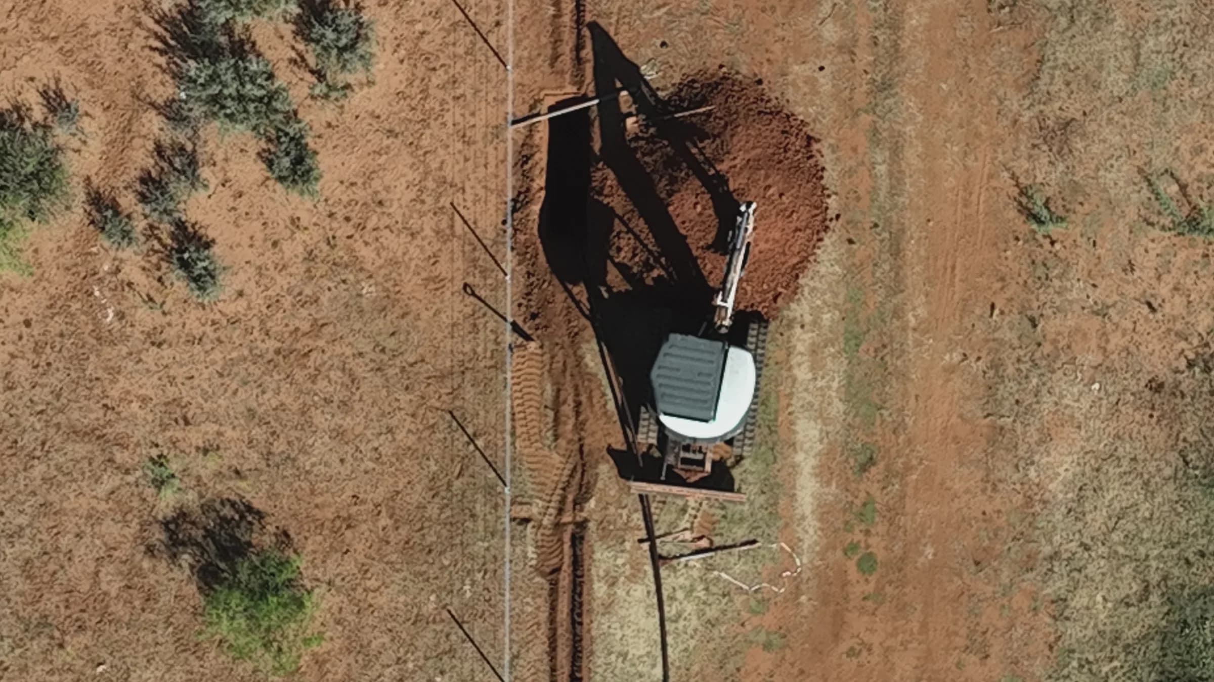 Overhead drone view of an excavator digging a pipeline trench along a fence line on red rangeland.