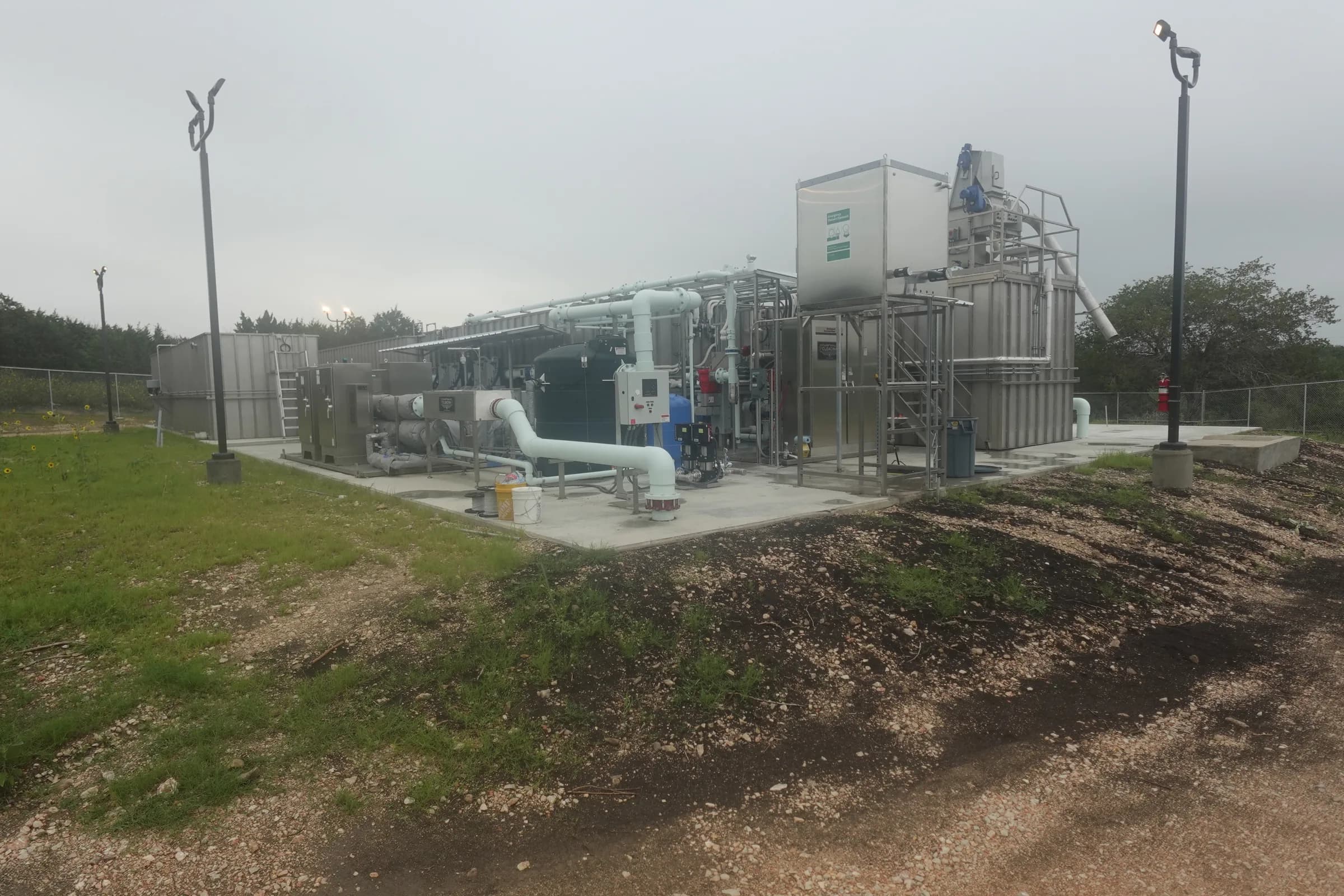 Packaged wastewater treatment plant on a concrete pad with white process piping, stainless steel tanks and site lighting under an overcast sky