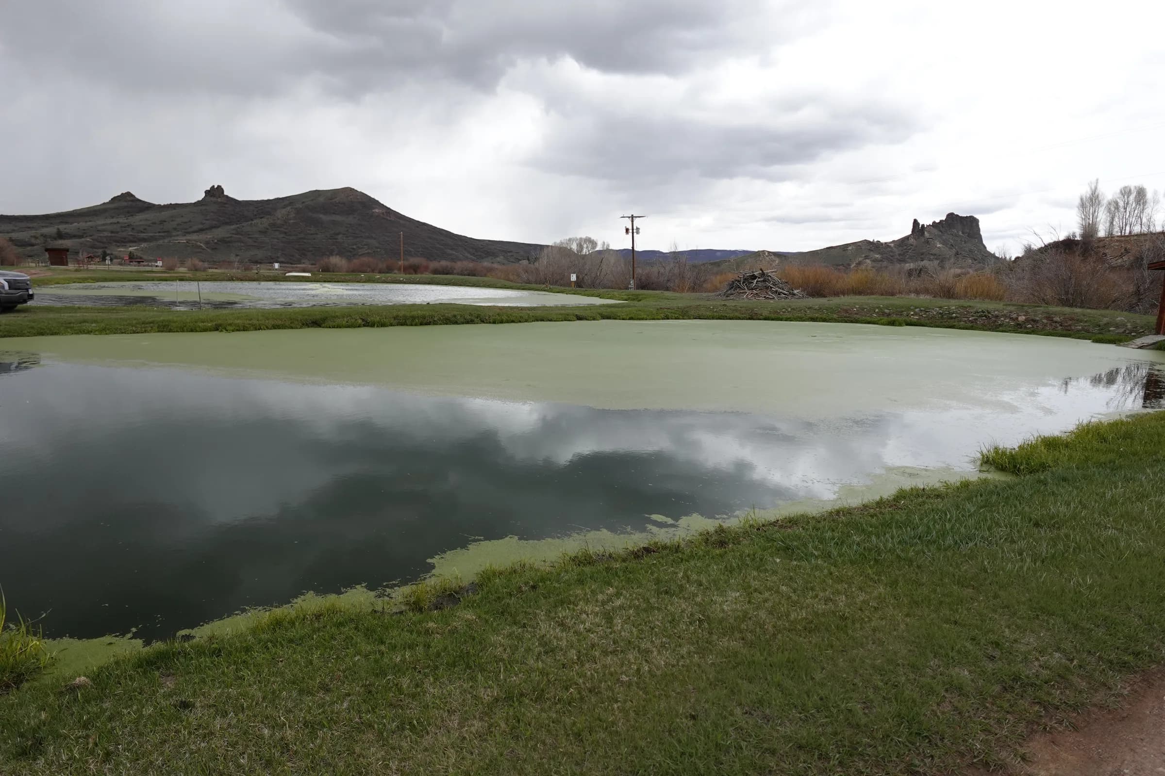 Two existing lagoon ponds with green algae on the surface in a grassy valley, with hills and a stormy sky behind