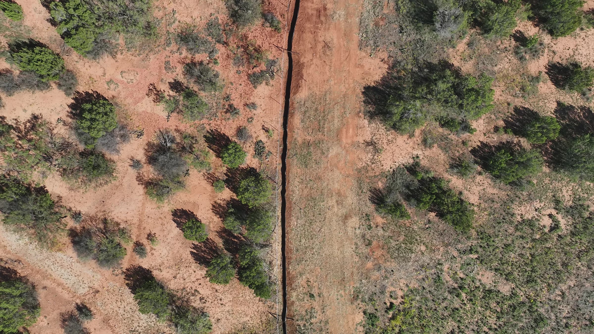 Drone view looking straight down on an open pipeline trench along a cleared corridor through red-soil scrubland