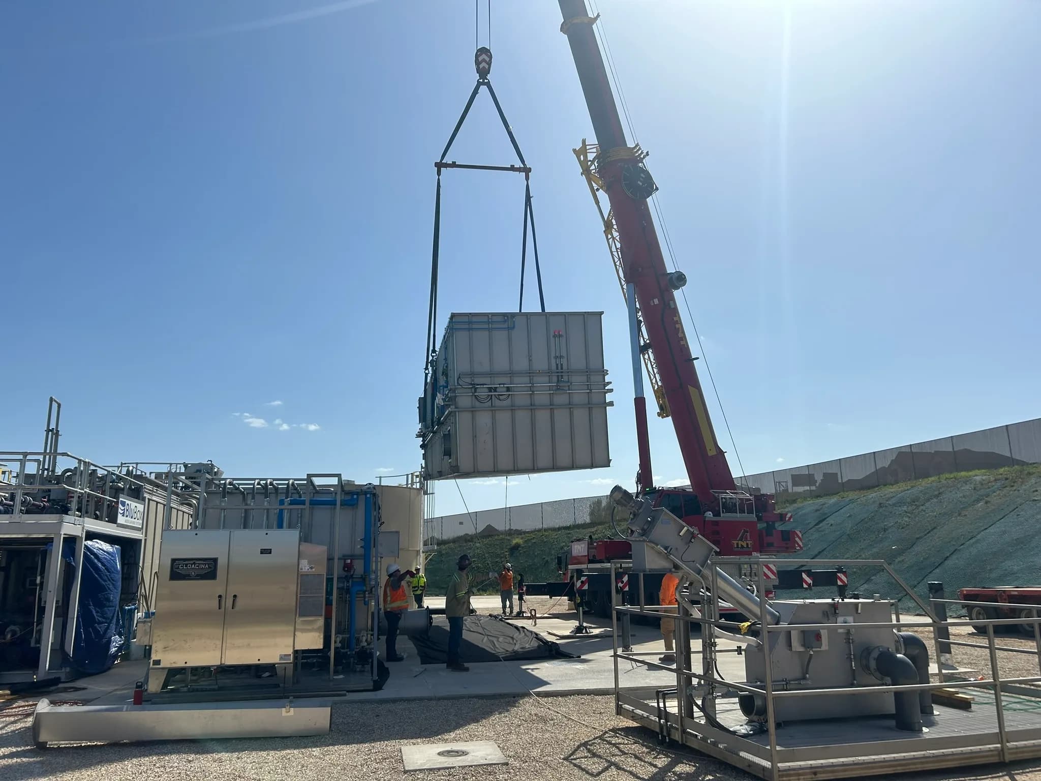 A mobile crane lifts a packaged wastewater treatment module into place beside other treatment units on a sunny day.