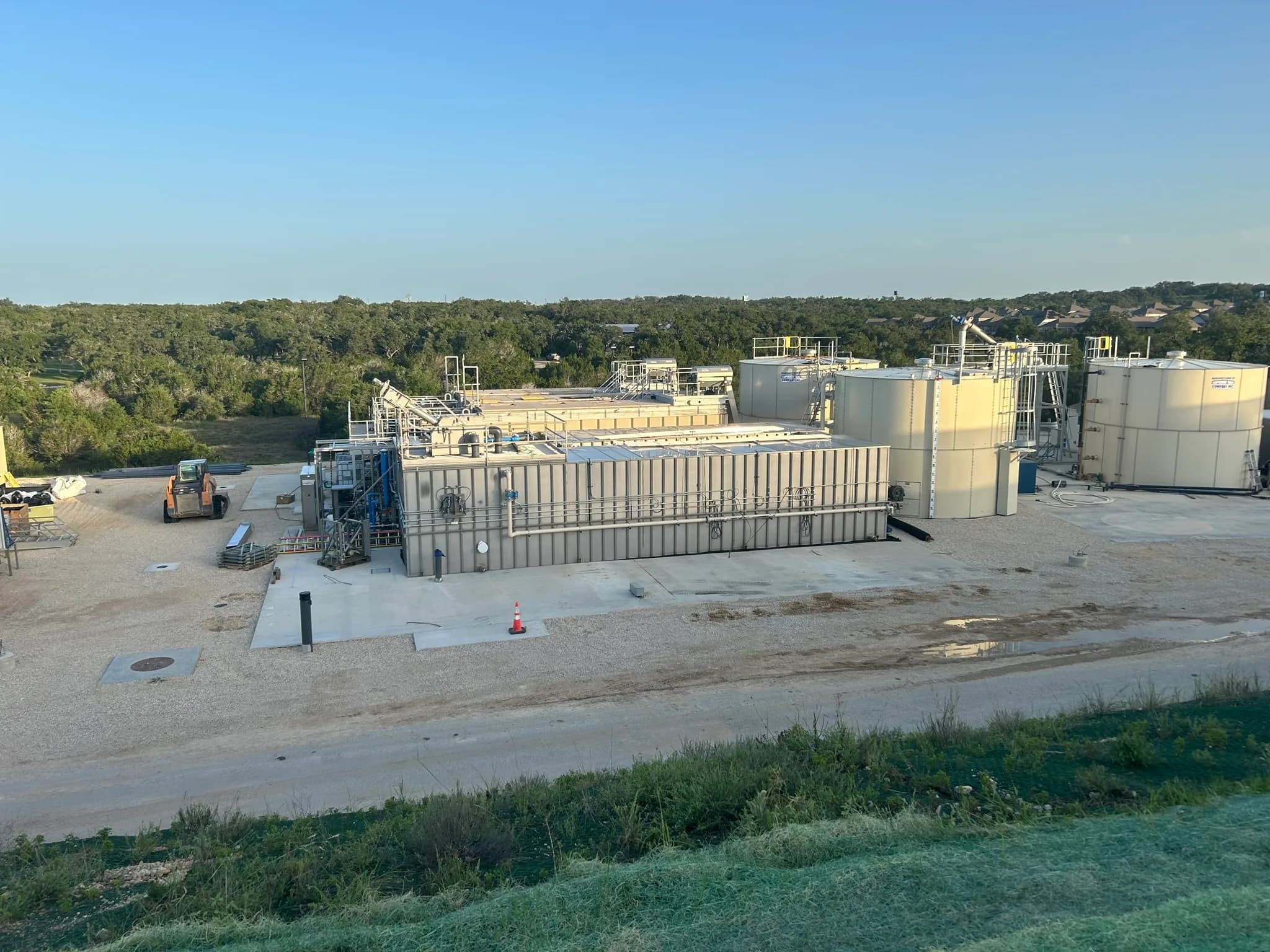 Packaged wastewater treatment plant seen from above, with cream-colored tanks, concrete pads and a skid steer.