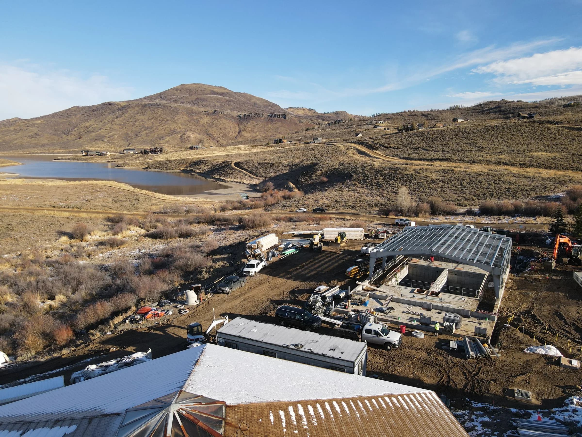 Drone view of a new wastewater treatment plant under construction, a steel frame over concrete basins on a graded site, with a reservoir and a mountain behind.
